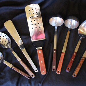 May include: A collection of stainless steel kitchen utensils with wooden handles. The set includes a slotted spatula, solid spatulas, ladles, and spoons. The handles are a dark brown color, and the metal parts are shiny.