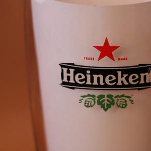 May include: A clear glass beer pint glass featuring the Heineken logo in black and white with a red star above the text. Below the brand name, there is a green hop illustration. The glass has a smooth, cylindrical shape and transparent walls.