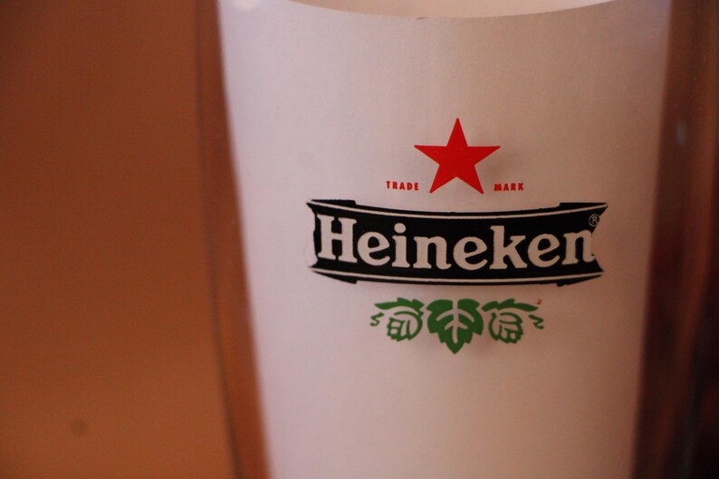 May include: A clear glass beer pint glass featuring the Heineken logo in black and white with a red star above the text. Below the brand name, there is a green hop illustration. The glass has a smooth, cylindrical shape and transparent walls.