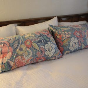 May include: Two decorative rectangular pillows with a floral pattern in shades of pink, blue, and yellow rest on a white bed. The pillows are placed on a white bedspread, with a wooden headboard in the background. The pillows add a touch of color to the bedroom.