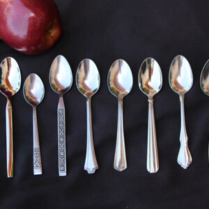 May include: Eight silver spoons of varying styles and designs are lined up on a black surface.