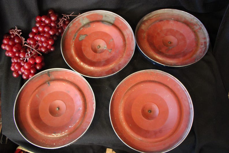 Puede incluir: Cuatro tapas met&aacute;licas rojas vintage con dise&ntilde;os de c&iacute;rculos conc&eacute;ntricos y bordes plateados est&aacute;n dispuestas sobre una superficie negra. Un racimo de uvas rojas est&aacute; en la esquina superior izquierda. Las tapas parecen ser de una olla o sart&eacute;n.