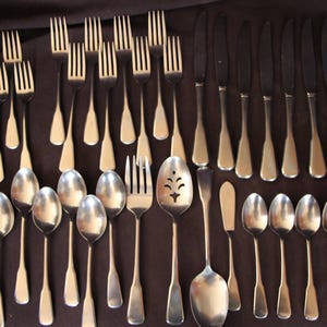 May include: A large collection of stainless steel flatware including various forks, knives, and spoons. The set features multiple dinner forks, salad forks, dinner knives, teaspoons, and soup spoons, along with a single slotted serving spoon.