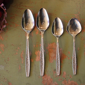May include: Four silver spoons of varying sizes are arranged on a distressed green surface. The spoons have textured handles and rounded bowls. A cluster of red grapes sits in a decorative metal basket in the upper left corner.