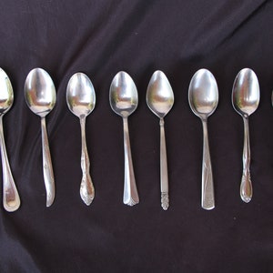 May include: Eight silver spoons of varying sizes and styles are lined up on a black surface. The spoons are all shiny and reflect light.