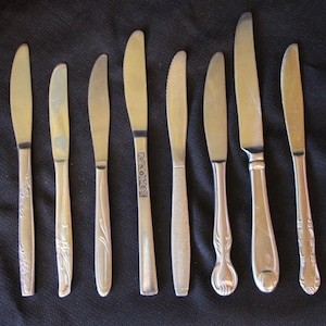 May include: An assortment of vintage butter knives with silver-colored blades and handles. The handles feature various decorative designs, including floral patterns and ornate shapes. The knives are arranged on a dark background.