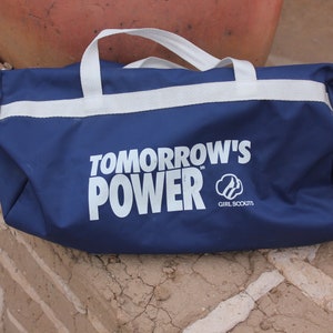 May include: A blue duffel bag with white straps and the text "TOMORROW'S POWER" in white letters. The Girl Scouts logo is also printed on the bag.