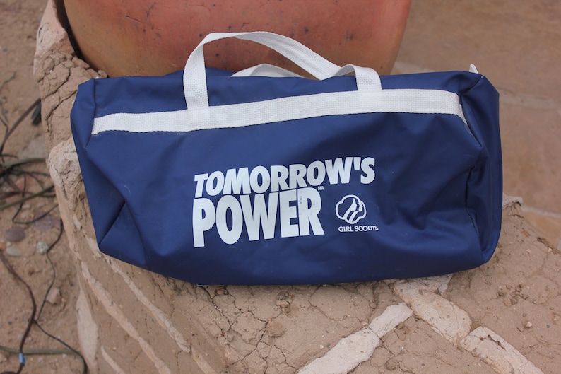 May include: A blue duffel bag with white straps and the text "TOMORROW'S POWER" in white letters. The Girl Scouts logo is also printed on the bag.