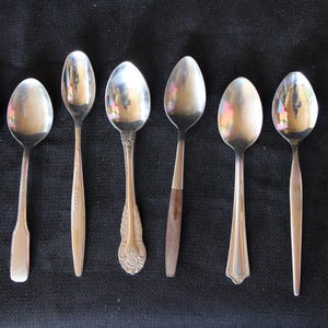 May include: Eight silver spoons with varying designs and finishes. The spoons are arranged in a row on a black surface.