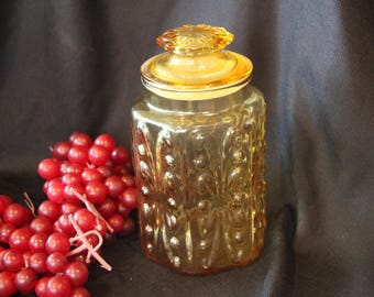 May include: A vintage amber glass jar with a decorative lid, featuring an embossed floral pattern. The jar is next to a cluster of red grapes, creating a still-life composition. The jar is likely used for storage.
