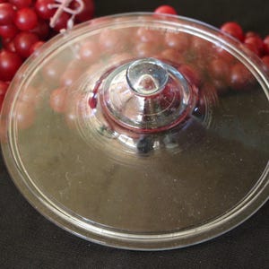 May include: A clear glass lid with a rounded edge and a central knob. The lid is circular and appears to be for a serving dish or bowl. Red grapes are visible in the background.