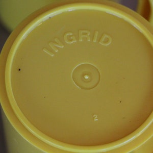 Op de afbeelding: Gele plastic beker met het woord "INGRID" erop gedrukt. De beker heeft een kleine cirkel in het midden en het nummer "2" eronder.