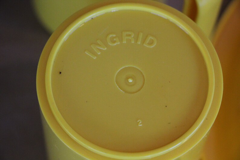 Op de afbeelding: Gele plastic beker met het woord "INGRID" erop gedrukt. De beker heeft een kleine cirkel in het midden en het nummer "2" eronder.
