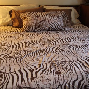 Puede incluir: Un juego de cama con un patr&oacute;n repetitivo de caras de cebra y tigre en marr&oacute;n y blanco en la funda n&oacute;rdica y una funda de almohada a juego. La ropa de cama est&aacute; dispuesta sobre una estructura de cama con almohadas lisas detr&aacute;s.
