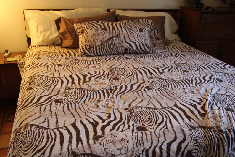 Puede incluir: Un juego de cama con un patr&oacute;n repetitivo de caras de cebra y tigre en marr&oacute;n y blanco en la funda n&oacute;rdica y una funda de almohada a juego. La ropa de cama est&aacute; dispuesta sobre una estructura de cama con almohadas lisas detr&aacute;s.