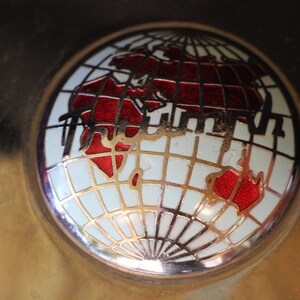 Puede incluir: Un emblema redondo y met&aacute;lico con un dise&ntilde;o de globo terr&aacute;queo. El globo muestra continentes rojos sobre un fondo blanco, con l&iacute;neas de cuadr&iacute;cula doradas. Los continentes est&aacute;n delineados en negro.