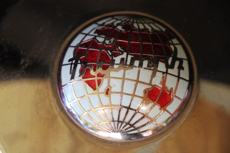 Puede incluir: Un emblema redondo y met&aacute;lico con un dise&ntilde;o de globo terr&aacute;queo. El globo muestra continentes rojos sobre un fondo blanco, con l&iacute;neas de cuadr&iacute;cula doradas. Los continentes est&aacute;n delineados en negro.