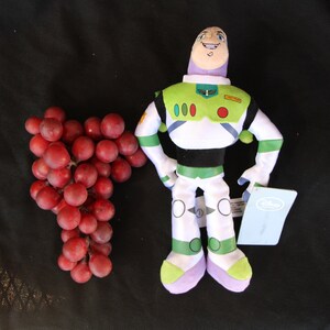 May include: A plush toy of Buzz Lightyear from the Toy Story franchise. The toy is wearing a green and white spacesuit with a red circle on the chest. The toy is standing on a black surface with a bunch of red grapes to the left.