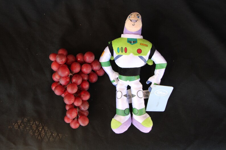 May include: A plush toy of Buzz Lightyear from the Toy Story franchise. The toy is wearing a green and white spacesuit with a red circle on the chest. The toy is standing on a black surface with a bunch of red grapes to the left.
