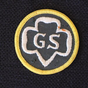 May include: A yellow and green Girl Scout patch with the letters "GS" embroidered in white.