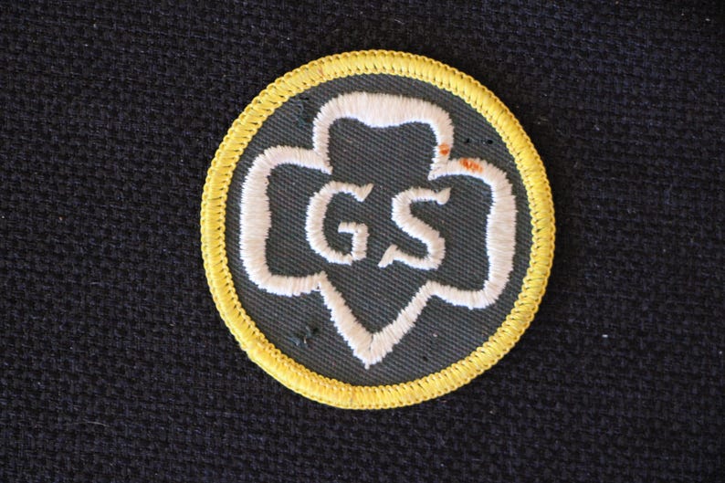 May include: A yellow and green Girl Scout patch with the letters "GS" embroidered in white.