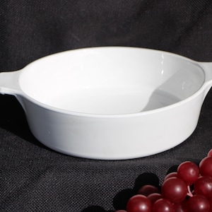 May include: A white, oval-shaped casserole dish with two small handles. The dish is empty and sits on a dark, textured surface. A cluster of red grapes is in the foreground.