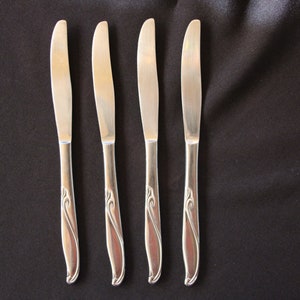 May include: Four silver butter knives with a curved handle design, arranged on a black surface. The knives have a simple, elegant design, with a smooth, polished finish. The handles feature a decorative, flowing pattern.
