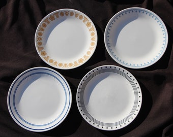 May include: Four white ceramic plates with different blue and gold rim designs. The plates are on a black fabric background.