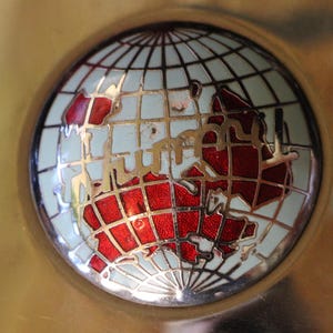 Puede incluir: Un objeto decorativo redondo de color dorado con un dise&ntilde;o detallado de globo terr&aacute;queo. El globo presenta un mapa del mundo rojo y blanco, sobre un fondo azul claro, y est&aacute; enmarcado por una cuadr&iacute;cula dorada.