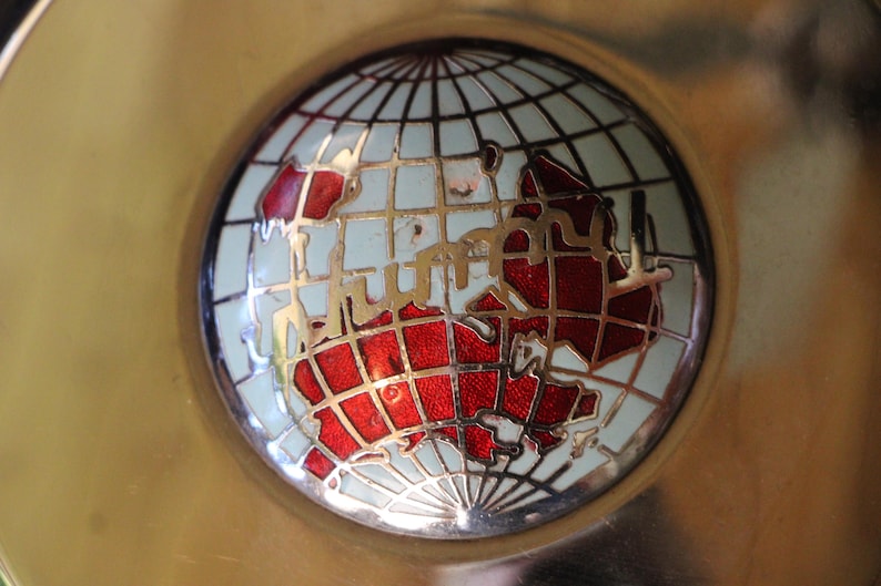 Puede incluir: Un objeto decorativo redondo de color dorado con un dise&ntilde;o detallado de globo terr&aacute;queo. El globo presenta un mapa del mundo rojo y blanco, sobre un fondo azul claro, y est&aacute; enmarcado por una cuadr&iacute;cula dorada.