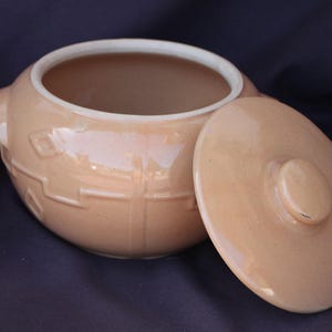 May include: A light peach-colored ceramic pot with a matching circular lid. The pot features a subtle embossed geometric pattern on its exterior surface and a smooth, glazed finish. It has a wide opening and a small handle on the side.