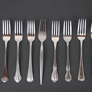 Puede incluir: Una colección de ocho tenedores de metal plateado dispuestos en una fila horizontal. Cada tenedor presenta un diseño de mango distinto, que varía desde formas simples y cónicas hasta patrones grabados ornamentados, todos con cuatro dientes y acabado pulido.