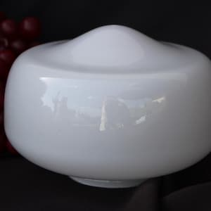 May include: A white, rounded glass lampshade with a smooth, glossy finish. The lampshade has a bulbous shape with a slightly raised top. The background is dark, with a cluster of red grapes to the left.