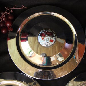 Puede incluir: Un conjunto de platos plateados vintage con un centro negro y un emblema decorativo con un mapa del mundo. Los platos tienen una superficie brillante y reflectante y est&aacute;n dispuestos sobre un fondo oscuro. Se ven uvas rojas.