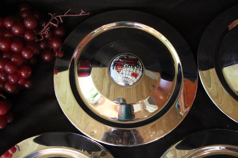 Puede incluir: Un conjunto de platos plateados vintage con un centro negro y un emblema decorativo con un mapa del mundo. Los platos tienen una superficie brillante y reflectante y est&aacute;n dispuestos sobre un fondo oscuro. Se ven uvas rojas.
