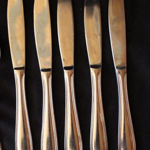 May include: A set of five silver-colored butter knives arranged vertically against a black background. Each knife has a rounded blade and a handle with a decorative, curved design. The knives are evenly spaced.