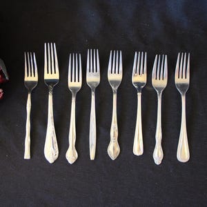 May include: Eight silver forks with different designs. The forks are arranged in a row on a black surface.