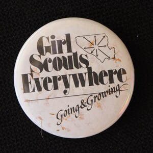 May include: White button with black text that reads "Girl Scouts Everywhere Going & Growing". The button features a black silhouette of a starburst shape.