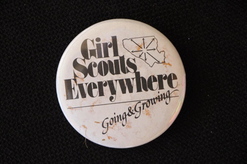 May include: White button with black text that reads "Girl Scouts Everywhere Going & Growing". The button features a black silhouette of a starburst shape.