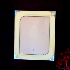 May include: A rectangular picture frame with a light yellow border and a central light tan insert. The frame features decorative silver-toned corner accents and thin horizontal lines at the top and bottom edges, creating a clean, structured appearance.