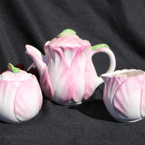 May include: A three-piece porcelain tea set with a floral design. The set includes a teapot, a sugar bowl with a lid, and a creamer. The pieces are white with pink petal-like designs and green accents.