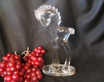 May include: A clear glass horse figurine with a detailed mane and tail stands on a round base. The horse is positioned next to a cluster of red grapes. The background is a dark, textured fabric.