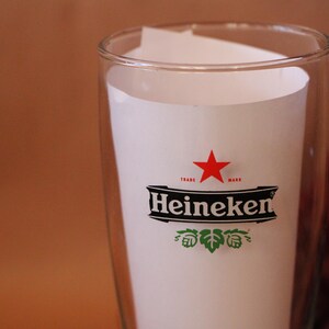 May include: A clear glass tumbler featuring the Heineken logo printed in black, white, and red. The logo includes a red star above the word Heineken in a bold black font, with green hop illustrations positioned directly below the brand name.