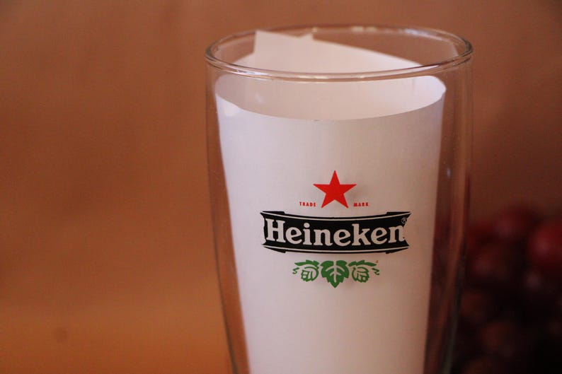 May include: A clear glass tumbler featuring the Heineken logo printed in black, white, and red. The logo includes a red star above the word Heineken in a bold black font, with green hop illustrations positioned directly below the brand name.