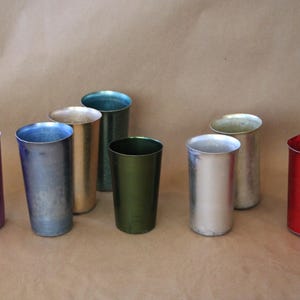 May include: An assortment of eight metallic drinking cups in various colors, including purple, blue, gold, green, silver, and red. The cups are cylindrical with a slightly flared rim and sit on a neutral background.