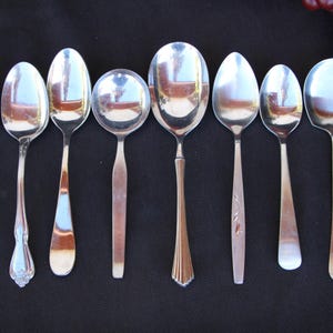 May include: A collection of eight silver spoons with varying handle designs. The spoons are arranged in a row, each reflecting the surrounding environment. The handles showcase different patterns and shapes, adding to the variety of the set.