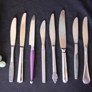 May include: An assortment of silver butter knives with various handle designs, including a purple handle, arranged on a dark surface. A bunch of green grapes is in the upper left corner.
