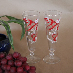 May include: Two clear glass beer glasses with red and white designs. The glasses have the words "Bière" and "Cervesa" written on them.