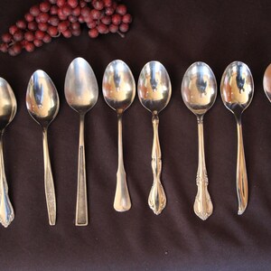 Puede incluir: Una colección de ocho cucharas de metal plateado dispuestas en una fila horizontal. Cada cuchara presenta un diseño de mango distinto, que varía desde estilos lisos y sencillos hasta patrones ornamentados y en relieve.