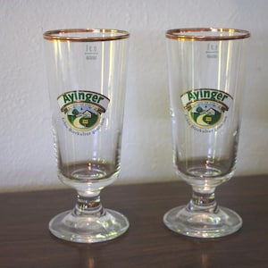 May include: Two clear Ayinger beer glasses with gold rims. Each glass has a logo with the word "Ayinger" and a landscape scene. The glasses are on a dark brown surface.